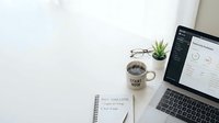 Modern workspace with laptop displaying analytics dashboard, coffee mug, succulent plant and notebook on clean white desk for productivity and business.