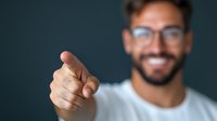 Young Hispanic businessman wearing glasses showing thumbs up gesture with confident smile against dark background. Professional corporate portrait.