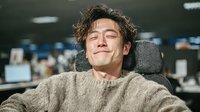 Young Asian male professional sleeping peacefully at office desk wearing cozy sweater in modern workplace environment for stress relief and work-life balance concepts.