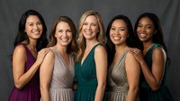 Five diverse women in elegant evening gowns smiling together against gray backdrop for formal event photography and celebration concepts.