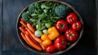 Fresh organic vegetables in rustic wooden bowl on dark background - carrots, tomatoes, bell peppers, broccoli and herbs arranged in colorful composition.