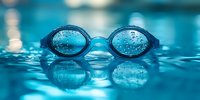 Swimming goggles with water drops floating on blue rippled water surface with reflection, dramatic sports equipment macro photography in pool atmosphere.