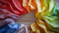 Vibrant rainbow colored silk fabric arranged in circular pattern on wooden surface creates soft flowing curves and folds. Luxurious textile background for design projects.