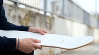 Person holding white skateboard deck outdoors with urban background, suitable for sports, lifestyle, and youth culture marketing materials.