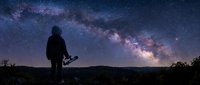 Silhouette of person with camera standing under spectacular Milky Way galaxy stretching across starry night sky over mountain landscape for astrophotography.