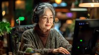 Senior Asian woman wearing headphones and glasses working at computer in evening office setting with warm lighting and bokeh background. Professional workspace atmosphere.