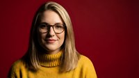 Professional blonde woman wearing glasses and yellow turtleneck sweater smiling confidently against red background for business and lifestyle concepts.