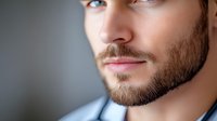 Close-up portrait of young Caucasian man with well-groomed beard and blue eyes against blurred background, showing confidence and masculinity in modern style.
