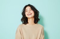 Young Asian woman with dark wavy hair laughing genuinely while wearing beige sweater against mint green background. Natural lifestyle portrait showing joy and happiness.