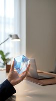 Person holding geometric crystal prism with blue and white facets at modern workspace with laptop, lamp, and potted plant by window with natural light.