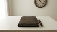Closed dark leather journal notebook with pen on white desk surface, wall clock in background, minimalist workspace setup for writing, planning, and organization.