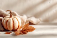 Small decorative white pumpkin with autumn leaves on beige fabric in soft sunlight, creating cozy fall atmosphere for seasonal decor.