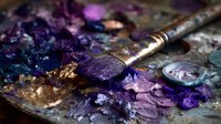 Artist's paintbrush with purple paint on a messy palette with vibrant purple, blue, and pink oil paint textures showing rich artistic process.