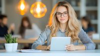 Young Caucasian businesswoman with blonde wavy hair and glasses working on digital tablet in modern coworking space, warm lighting and office background.
