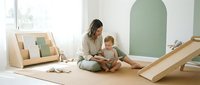 Young Caucasian mother sitting on floor reading book with toddler in bright Montessori playroom with wooden toys and natural decor.