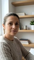 Middle-aged Hispanic woman in cozy sweater smiling warmly at home office with wooden shelves and books in background for lifestyle content.