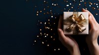 Female hands holding elegant gift box wrapped in silver paper with golden bow against dark background with festive bokeh lights and copy space for holiday season.
