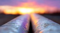Dramatic sunset reflection on metal pipeline extending to horizon, with vibrant orange sky and purple clouds creating atmospheric industrial landscape.