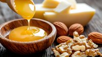 Golden honey dripping into wooden bowl with mixed nuts scattered on rustic wooden table, natural food photography with warm lighting and selective focus.