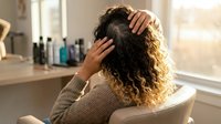 Young woman with curly hair touching head in bright bathroom, suggesting headache or stress relief moment during morning routine.