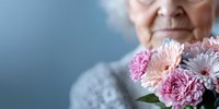 Senior Caucasian woman in white lace holding fresh pink and white flowers against blue background, cropped view showing gentle smile and bouquet arrangement.