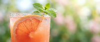 Refreshing grapefruit cocktail with ice and fresh mint sprig in glass, set against soft green bokeh background on sunny day.