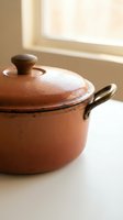 Vintage terracotta cooking pot with lid and metal handles sits on white surface near bright window, showcasing rustic kitchenware and nostalgic home cooking heritage.