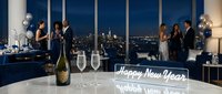 Elegant New Year Eve rooftop party with diverse guests, champagne bottle, glowing Happy New Year sign, city skyline at night, blue decor.