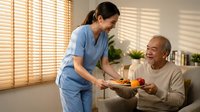 Asian female nurse in blue scrubs serving healthy meal to smiling elderly Caucasian man in bright home care setting with natural window light.
