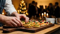 Chef preparing gourmet holiday appetizers on wooden board with Christmas tree lights and dinner party guests in warm restaurant setting.