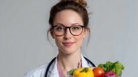 Young female nutritionist in white medical coat and glasses holding fresh vegetables and fruits, promoting healthy eating habits and balanced diet.