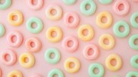 Colorful pastel cereal rings scattered on pink background in flat lay composition, creating playful breakfast pattern for food and confectionery design projects.