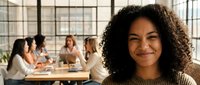 Smiling mixed-race businesswoman with curly hair in modern office workspace with diverse team meeting in background for corporate and professional content.