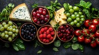 Fresh berries, grapes, tomatoes and cheese assortment in ceramic bowls on dark background with herbs and leaves, top view composition for gourmet appetizer.
