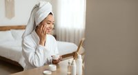 Young Asian woman in white bathrobe and towel applying skincare cream while looking in mirror during morning beauty routine in modern bedroom.