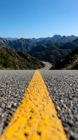 Winding mountain road with yellow dividing line stretching into rugged mountain range under clear blue sky, low angle view focusing on road marking.