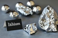 Raw lithium metal pieces with silvery metallic luster on dark surface near identification label, showing pure element specimens for scientific or educational purposes.