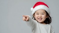 Cheerful child wearing Santa hat pointing with excitement against neutral gray background, perfect for holiday season advertising.