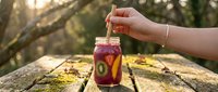 Woman holding wooden spoon over jar of homemade fruit preserve or jam outdoors on rustic wooden surface with forest background, food preparation and preservation.