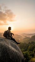 Male hiker with backpack sitting on rocky boulder overlooking misty forested valley at golden sunrise, contemplating scenic mountain landscape for travel and adventure concepts.