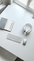 Clean white minimalist workspace with wireless keyboard, mouse, headphones and notebook on desk near window for productivity and remote work.