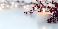 Burning white candle with red winter berries and soft bokeh lights creating cozy holiday atmosphere on light background. Perfect for seasonal greetings.