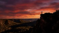Silhouetted figure standing on rocky cliff edge overlooking vast mountain valley at dramatic sunset with fiery orange and purple sky, conveying adventure and exploration themes for travel and lifestyle content.