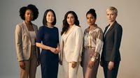 Diverse group of five professional women standing together with joined hands against neutral background, representing unity, teamwork, collaboration, and corporate diversity in business environment.