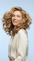 Happy young Caucasian woman with flowing blonde curly hair in motion wearing cream silk blouse against light blue background for beauty and lifestyle concepts.