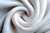 Soft white knitted fabric swirled in spiral pattern creating textural waves and folds, macro close-up showing detailed textile weave structure and natural light shadows.