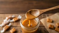 Golden honey dripping from wooden spoon into glass jar on rustic wood background with caramel candies for natural sweetness and culinary applications.