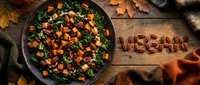 Autumn kale salad with roasted sweet potato, pecans, and cranberries on rustic wood, with the word vegan spelled in nuts beside fall leaves.