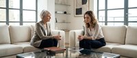 Professional mature Caucasian female therapist counseling young Hispanic woman client during therapy session in modern bright office with white sofa.