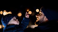 Young African American boy points at night sky while bearded Caucasian father watches with joy, bokeh lights create magical winter atmosphere against dark background.